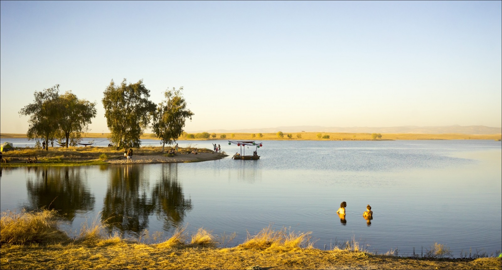 Why Humans Need Festivals 1 Two festivalgoers stand in a lake near a tree-lined shore at sunset.