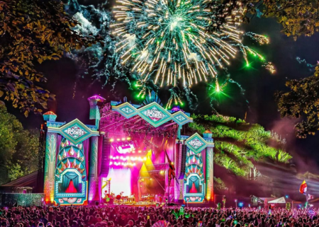 Colorful Electric Forest festival scene with lights, stage design, and people moving through the grounds.